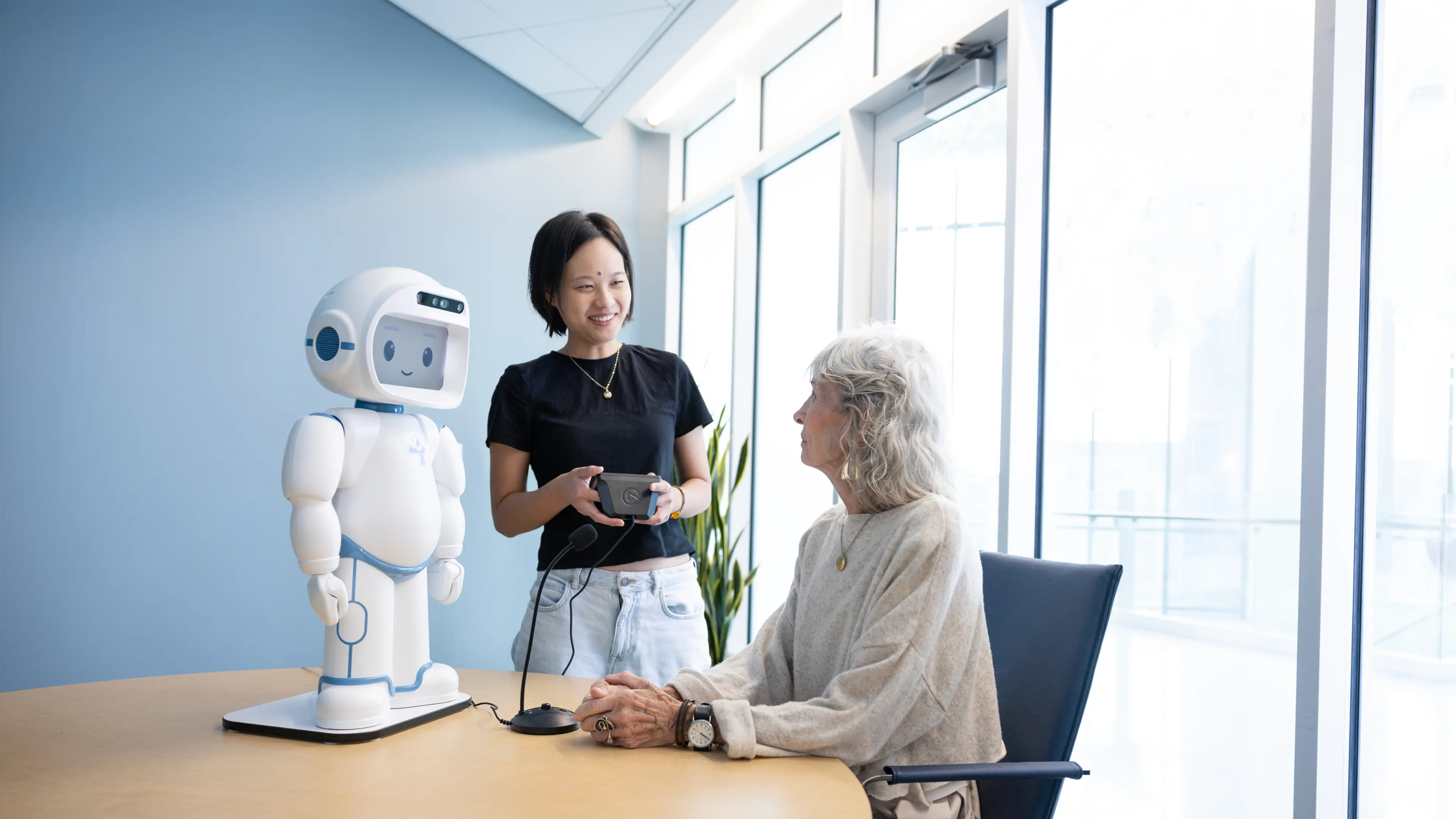 A female student stands next to a table where an older woman sits; a white robot stands on the table in front of them both.