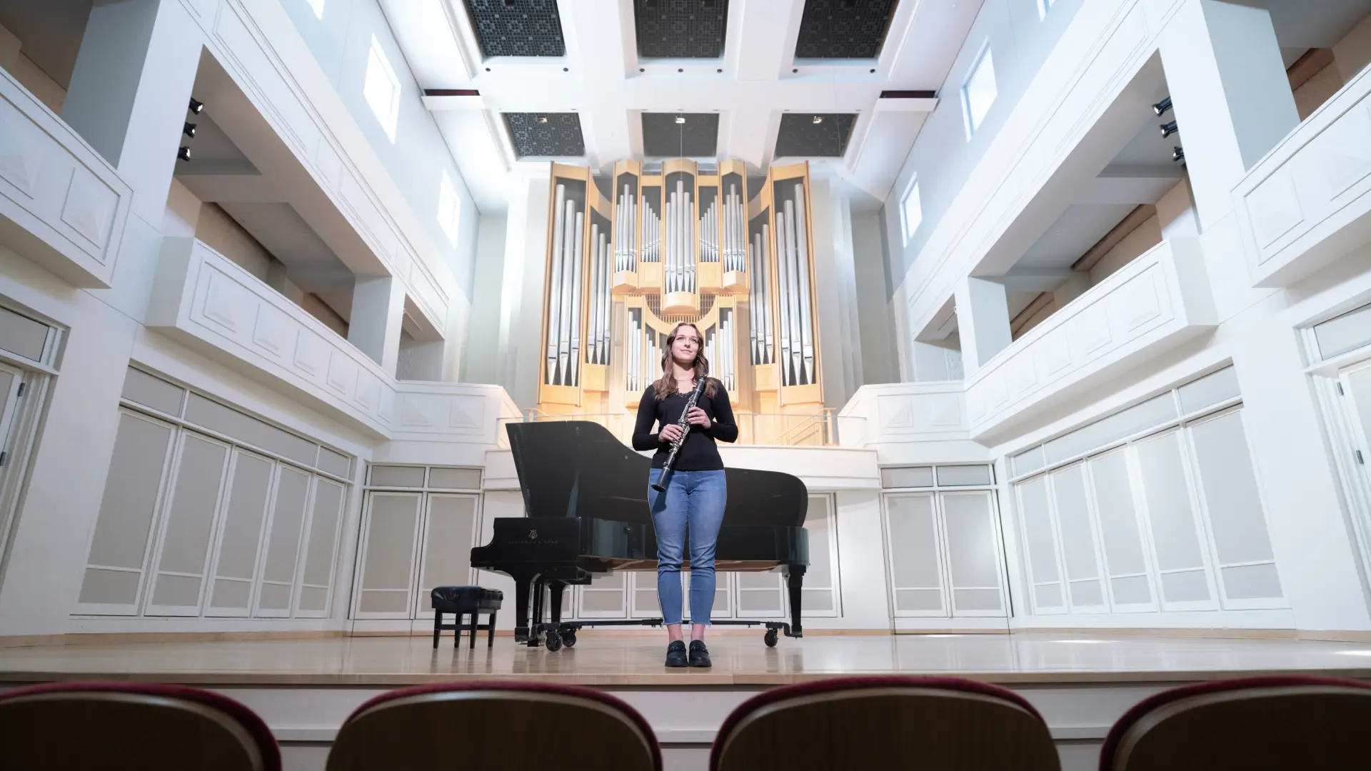A female student stands centerstage holding her instrument, she looks out across the performance hall in front of her.