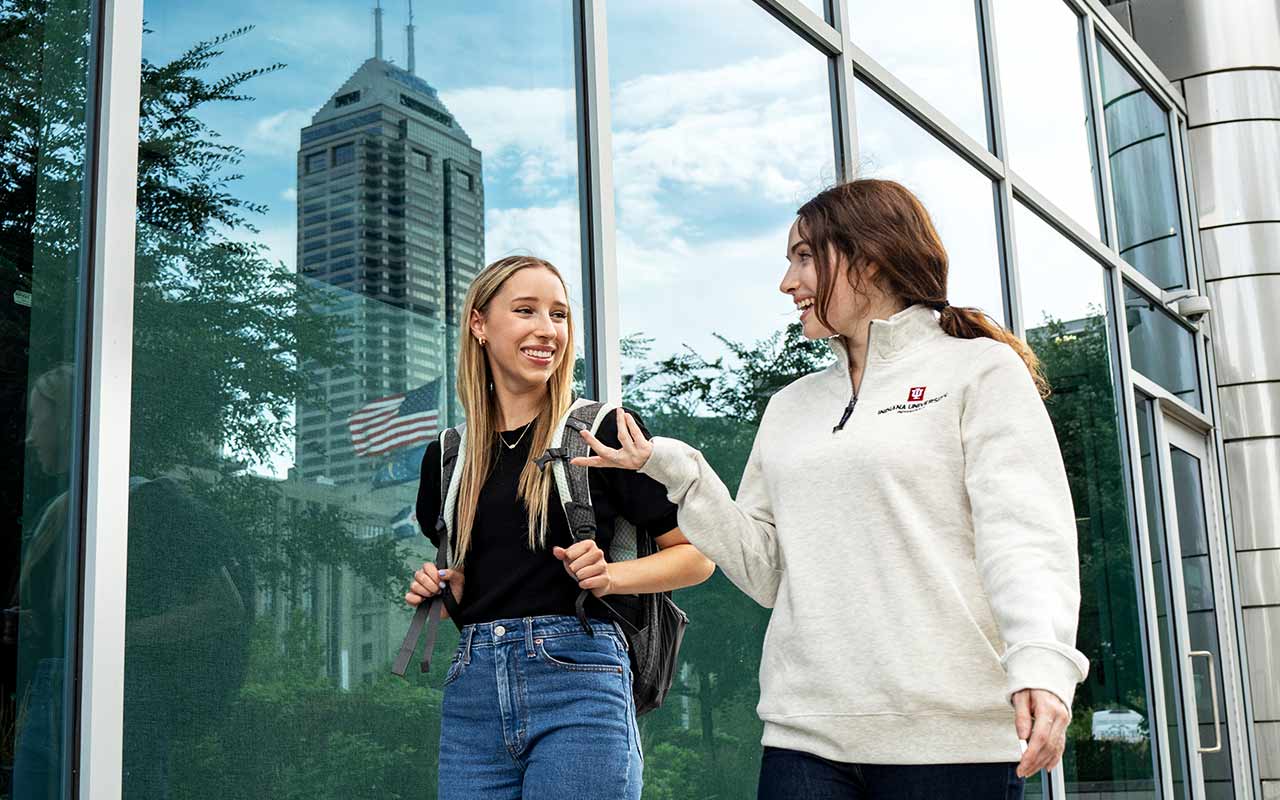 Two female students talk while walking down a city street, the reflection of Salesforce tower stands next to them in a glass window.