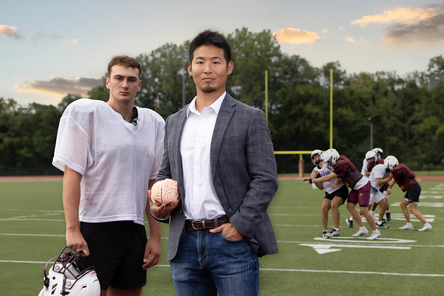 Kei Kawata stands with a high school football player while holding a brain during a scrimmage on a football field