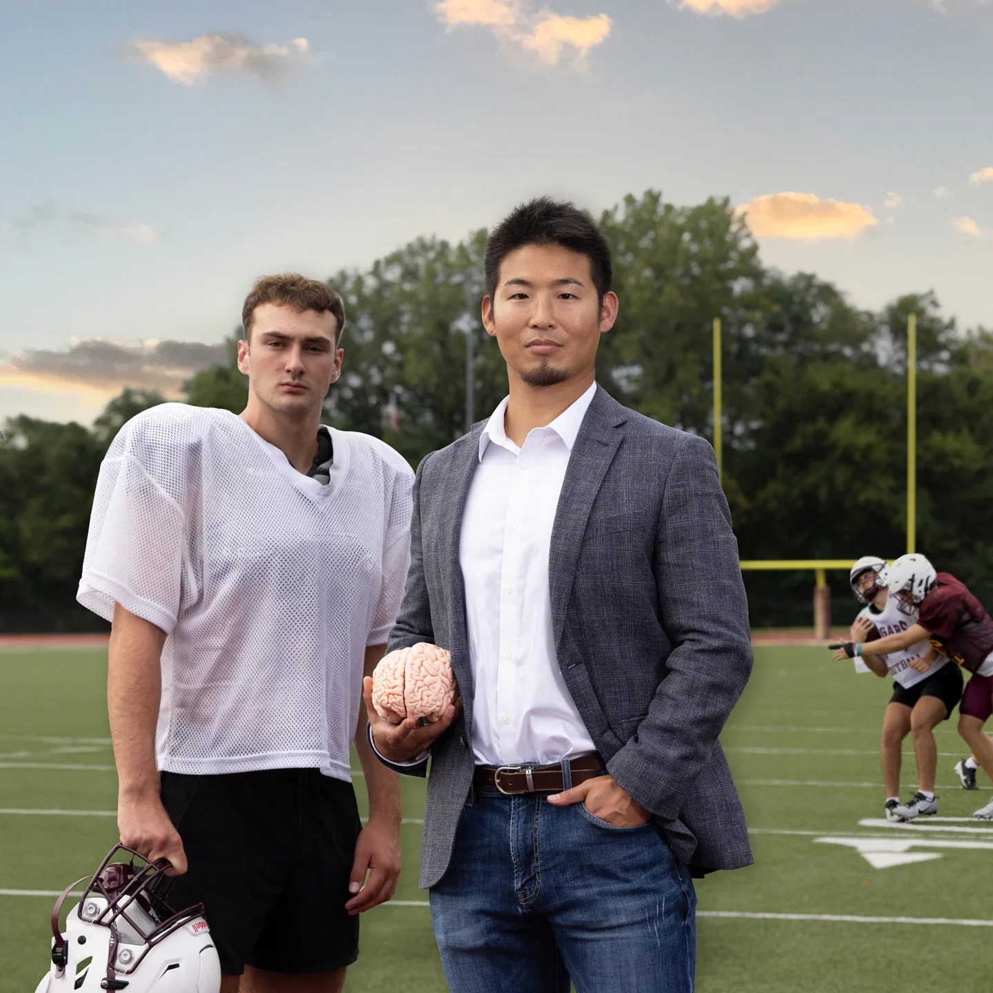 Kei Kawata stands with a high school player on a football field holding a brain while a scrimmage takes place in the background. 