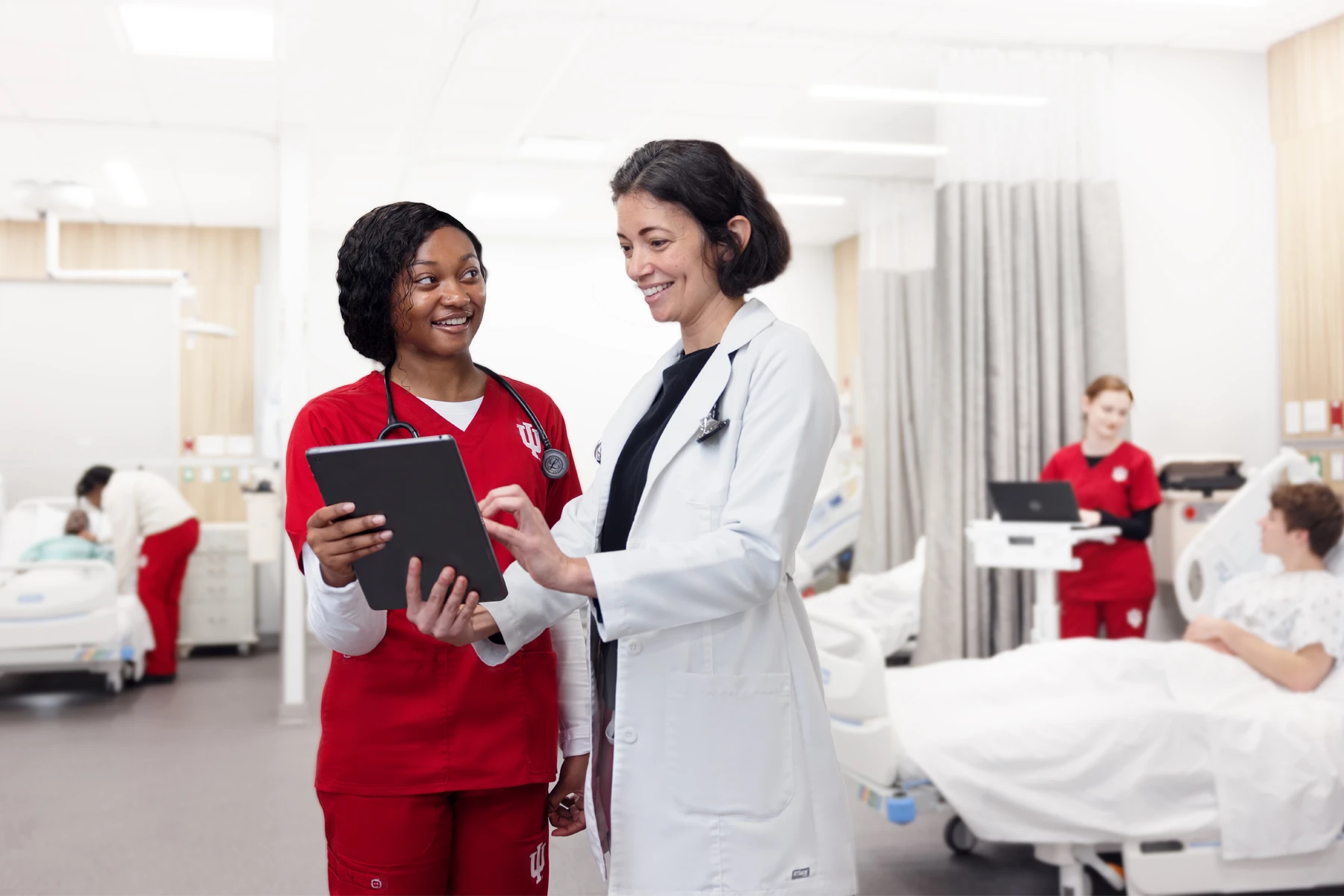 A nursing student and instructor review a chart on a tablet while other students work in the simulation lab behind them.