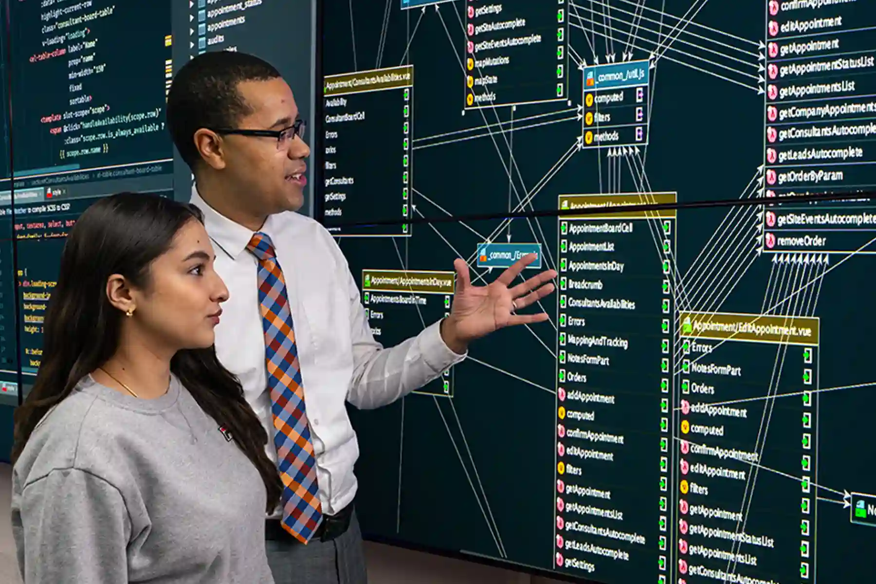 A male professor explains a large screen of cybersecurity flowcharts to a female student, listening intently.