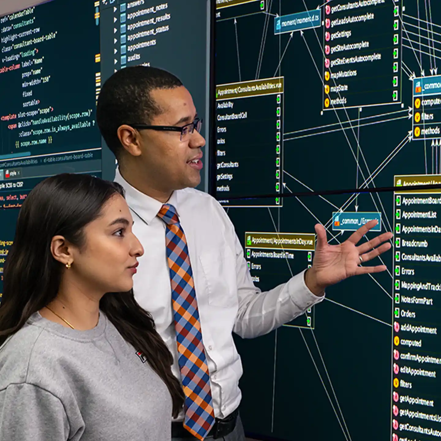 A male professor explains a large screen of cybersecurity flowcharts to a female student, listening intently.