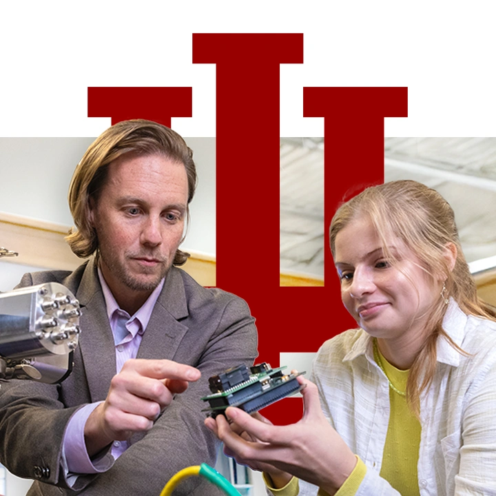 A male instructor and a female student stand behind a table filled with electronics equipment. She holds a microelectronic device and the instructor is pointing to it. A large trident is layered behind the subjects.