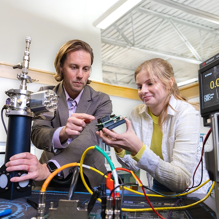 A male instructor and a female student stand behind a table filled with electronics equipment. She holds a microelectronic device and the instructor is pointing to it