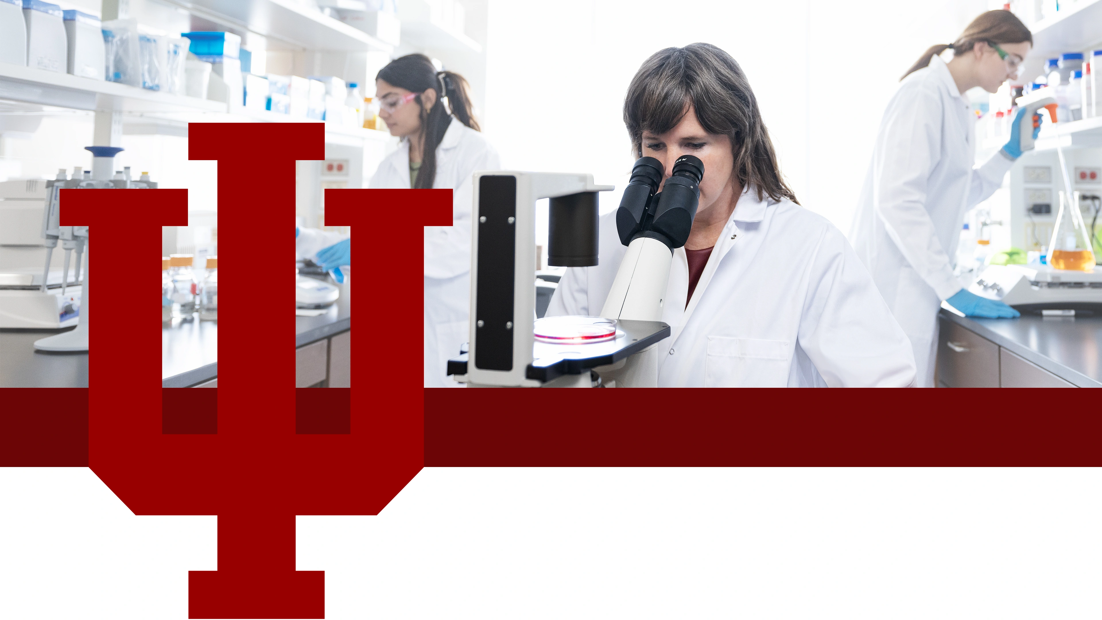 Three female researchers in lab coats work in a bright laboratory. A large red IU trident is overlaid.