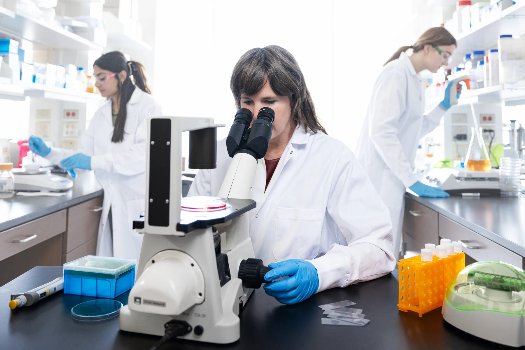 A scientist looks into a microscope while 2 student researchers work in the lab behind her.