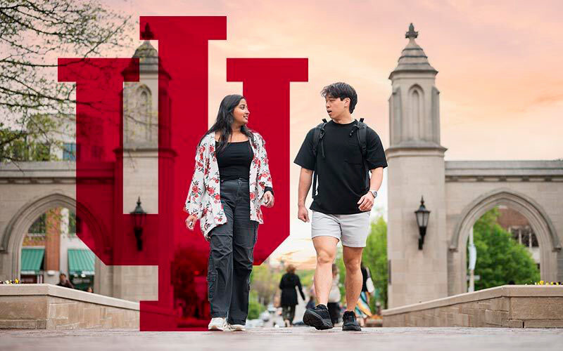 Two IU Bloomington students walk in front of the Sample Gates. A crimson IU trident is in the background.