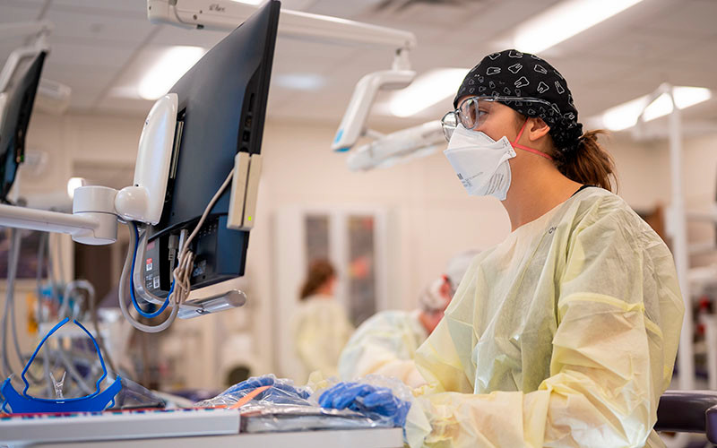 An IU Fort Wayne dentistry student wearing scrubs and a mask works at a computer.