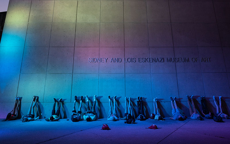 The Light Totem covers the Sidney and Lois Eskenazi Museum of Art in a rainbow of colors as IU students lie on their backs with their feet up on the wall.