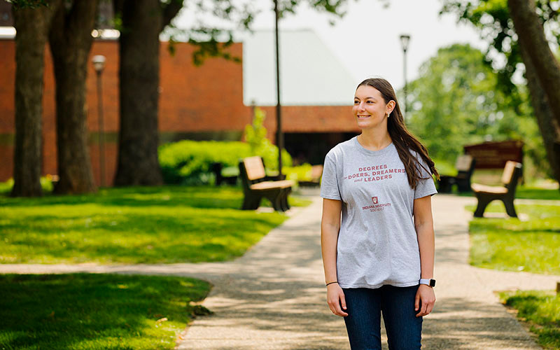 A student stands in a green space on the IU Southeast campus. She wears an IUS shirt that says, ‘Degrees for doers, dreamers, and leaders.’