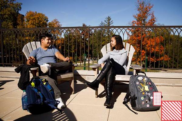Two students sit on an outdoor patio having a conversation.